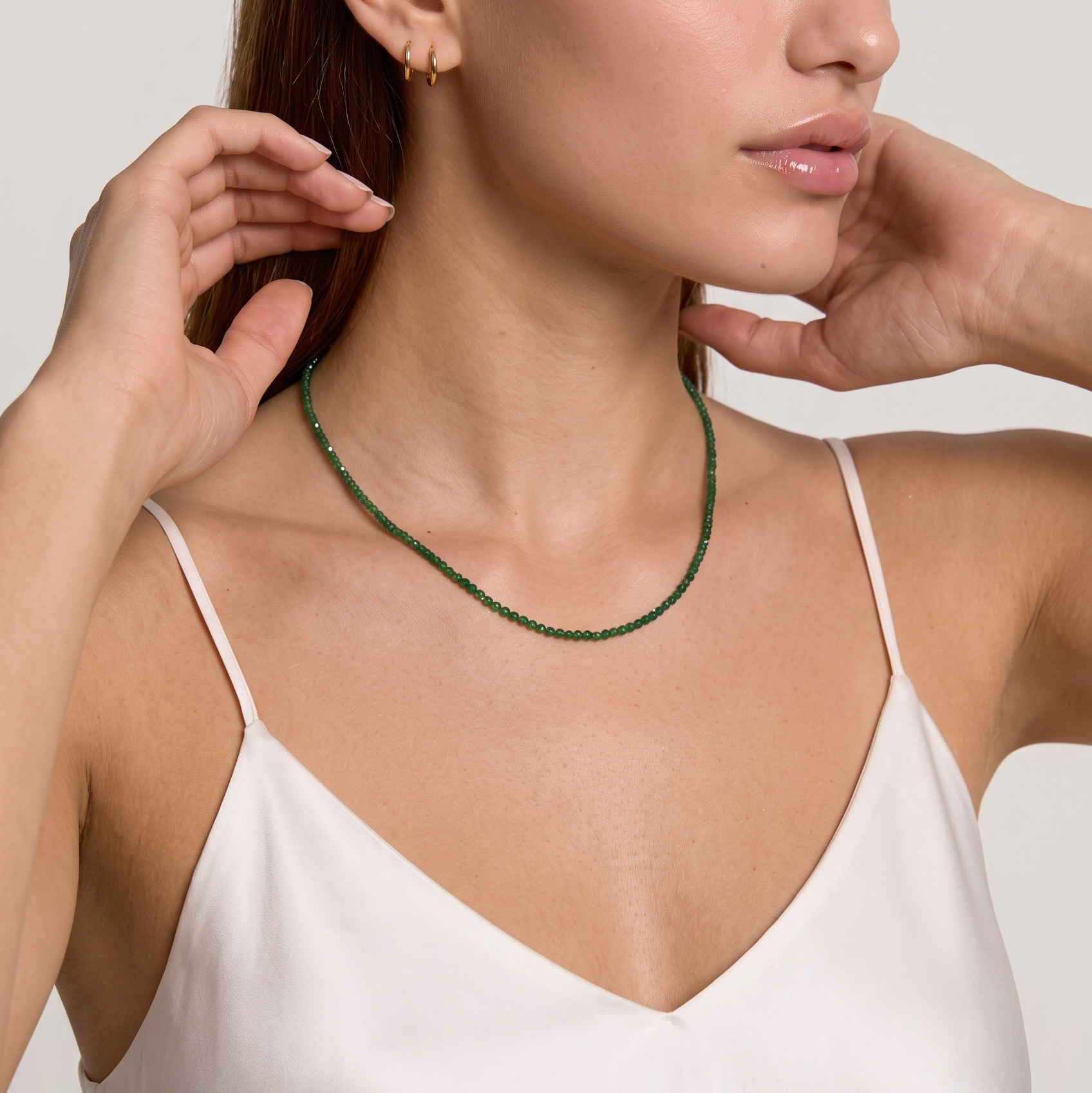 A woman wearing a delicate green beaded necklace and a small gold hoop earring, posing in a white camisole against a light gray studio background.