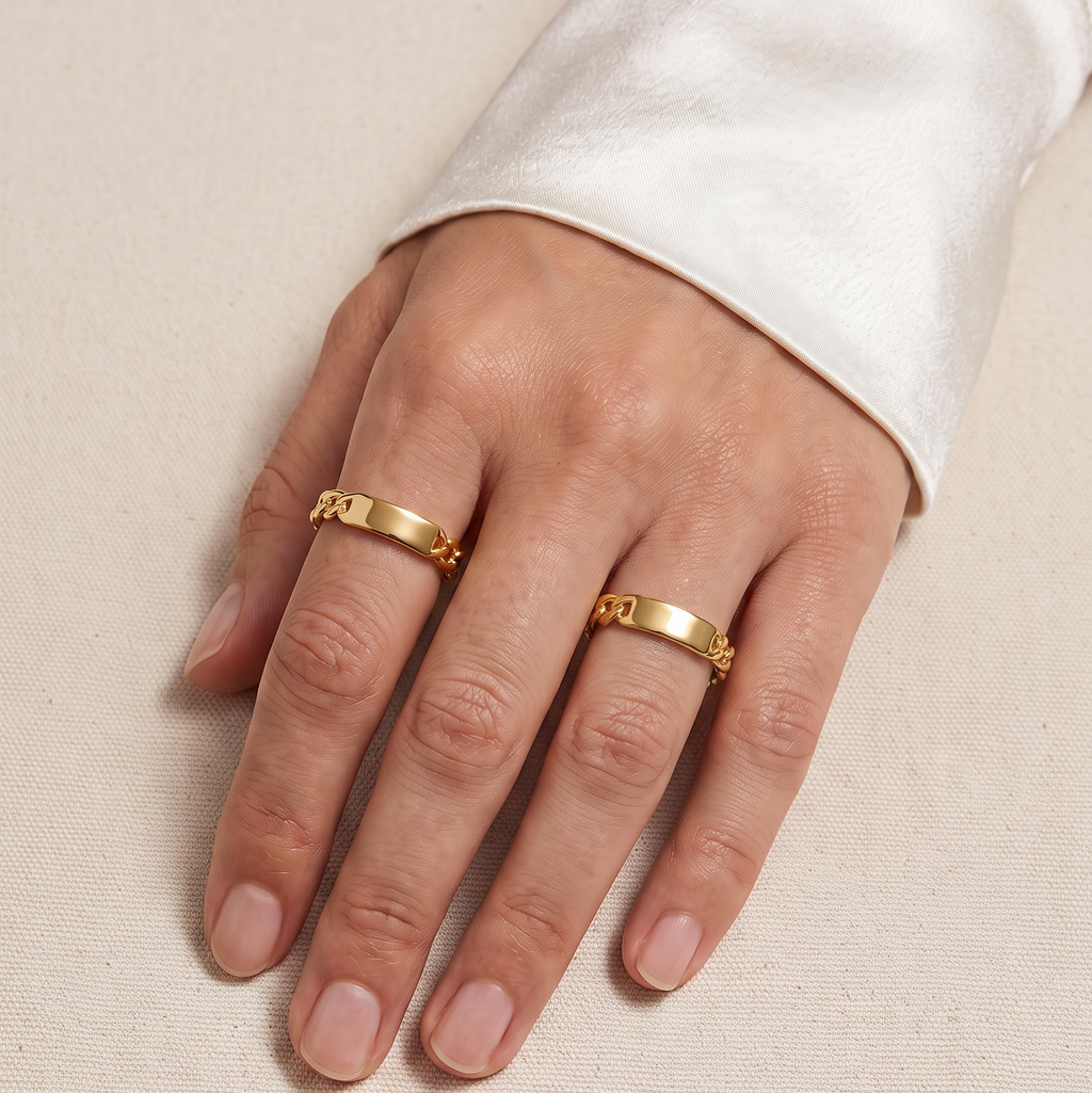 A close-up of a hand wearing two gold rings with engraved lettering, styled with a white satin sleeve against a neutral beige fabric background.
