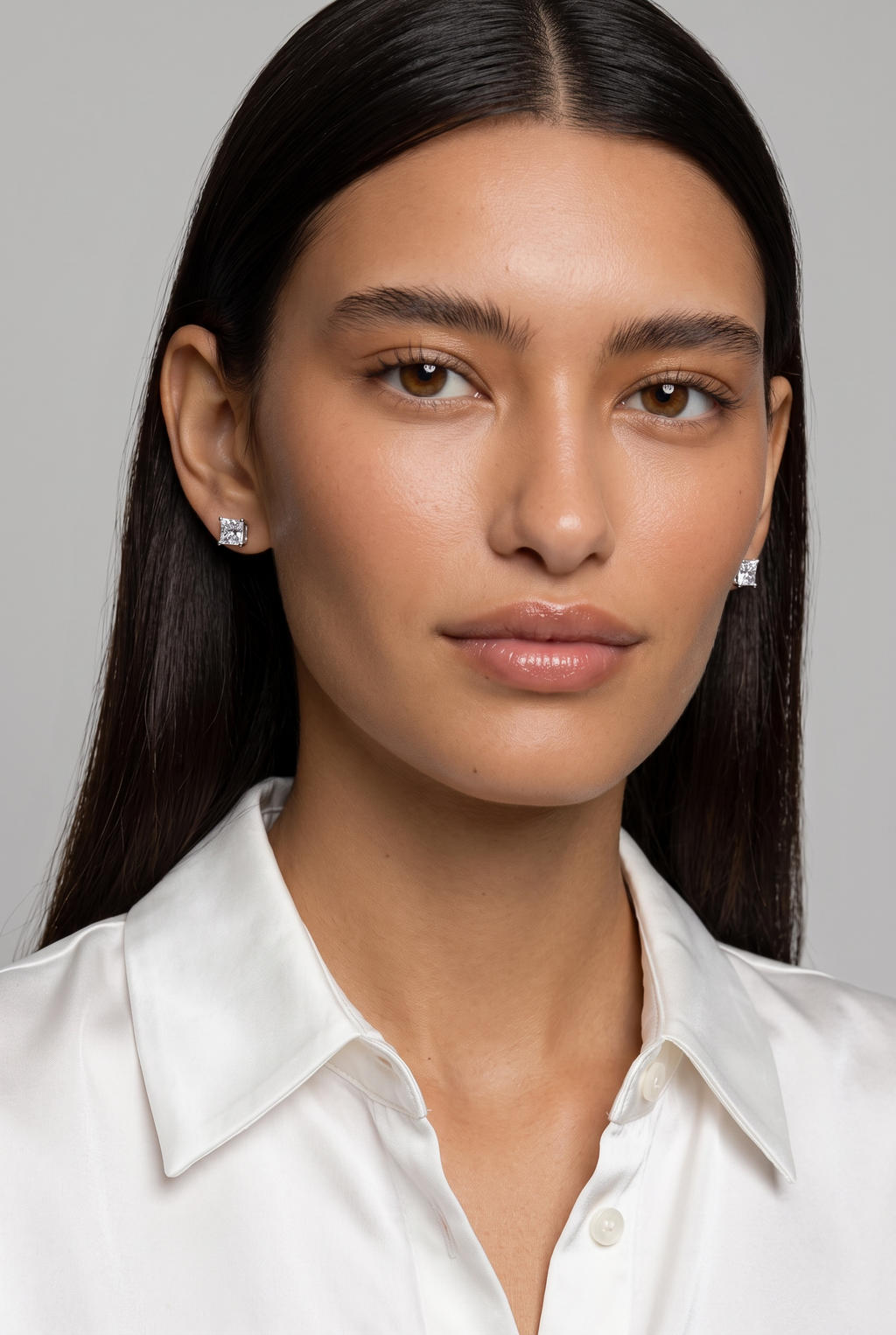 A close-up portrait of a woman with long dark hair wearing small silver stud earrings and a white collared shirt against a light gray background.