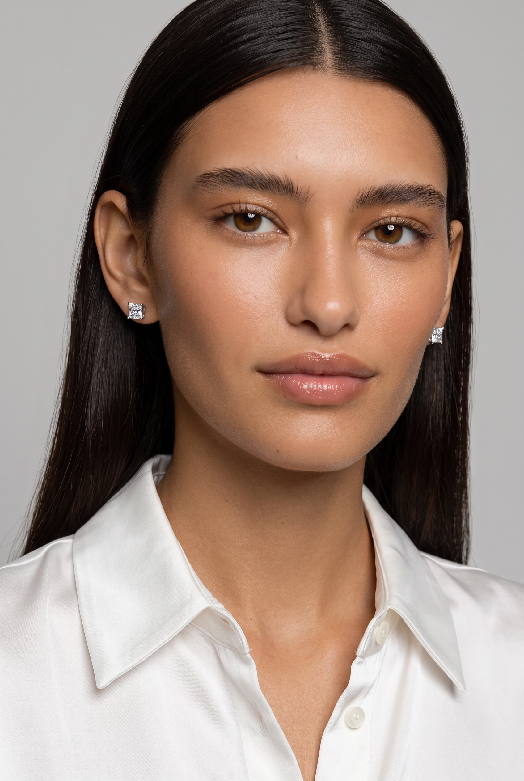 A close-up portrait of a woman with long dark hair wearing small silver stud earrings and a white collared shirt against a light gray background.