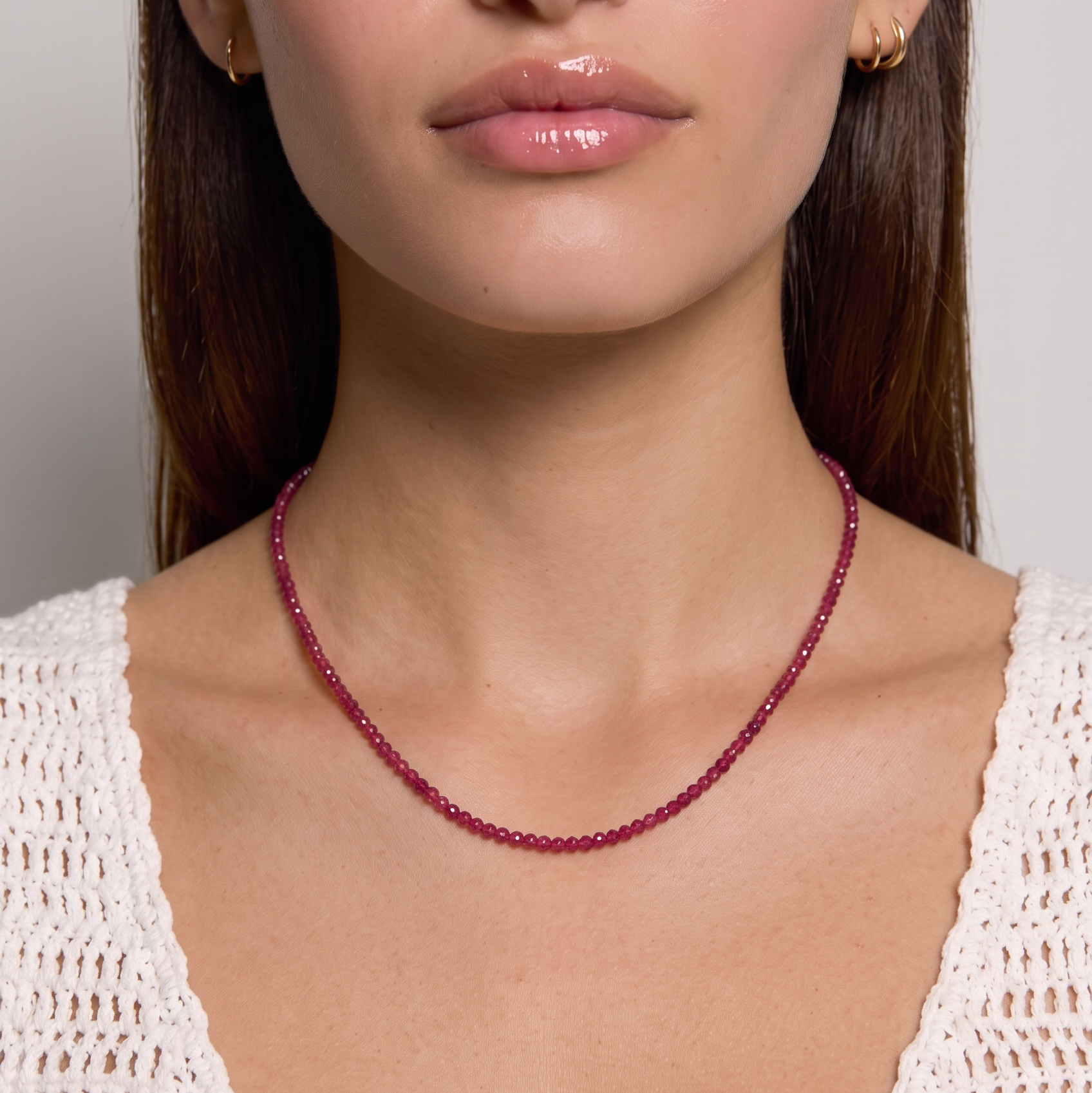 A close-up of a woman wearing a delicate, thin chain necklace made of small dark red beads, paired with a white knit top against a neutral gray background.