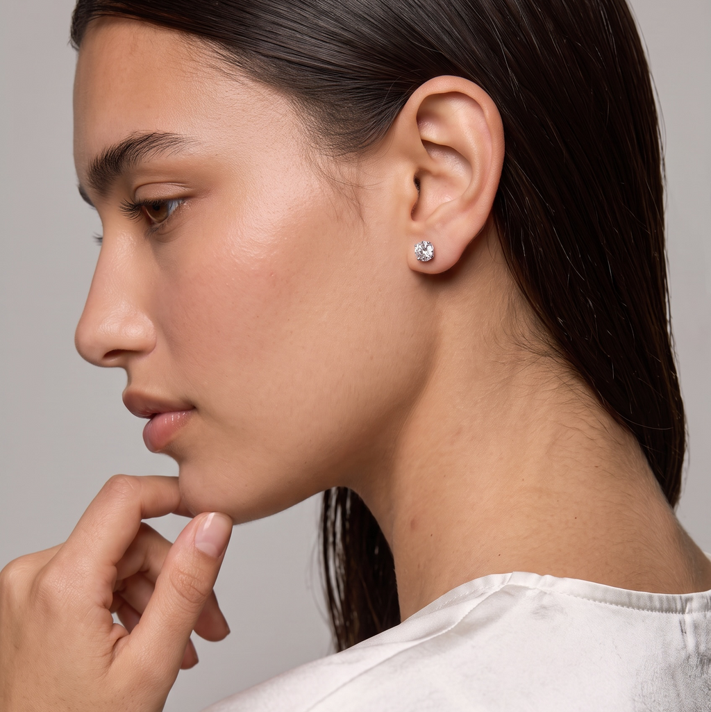 A close-up side profile of a woman wearing a small round clear gemstone stud earring, dressed in a white satin top against a neutral gray background.