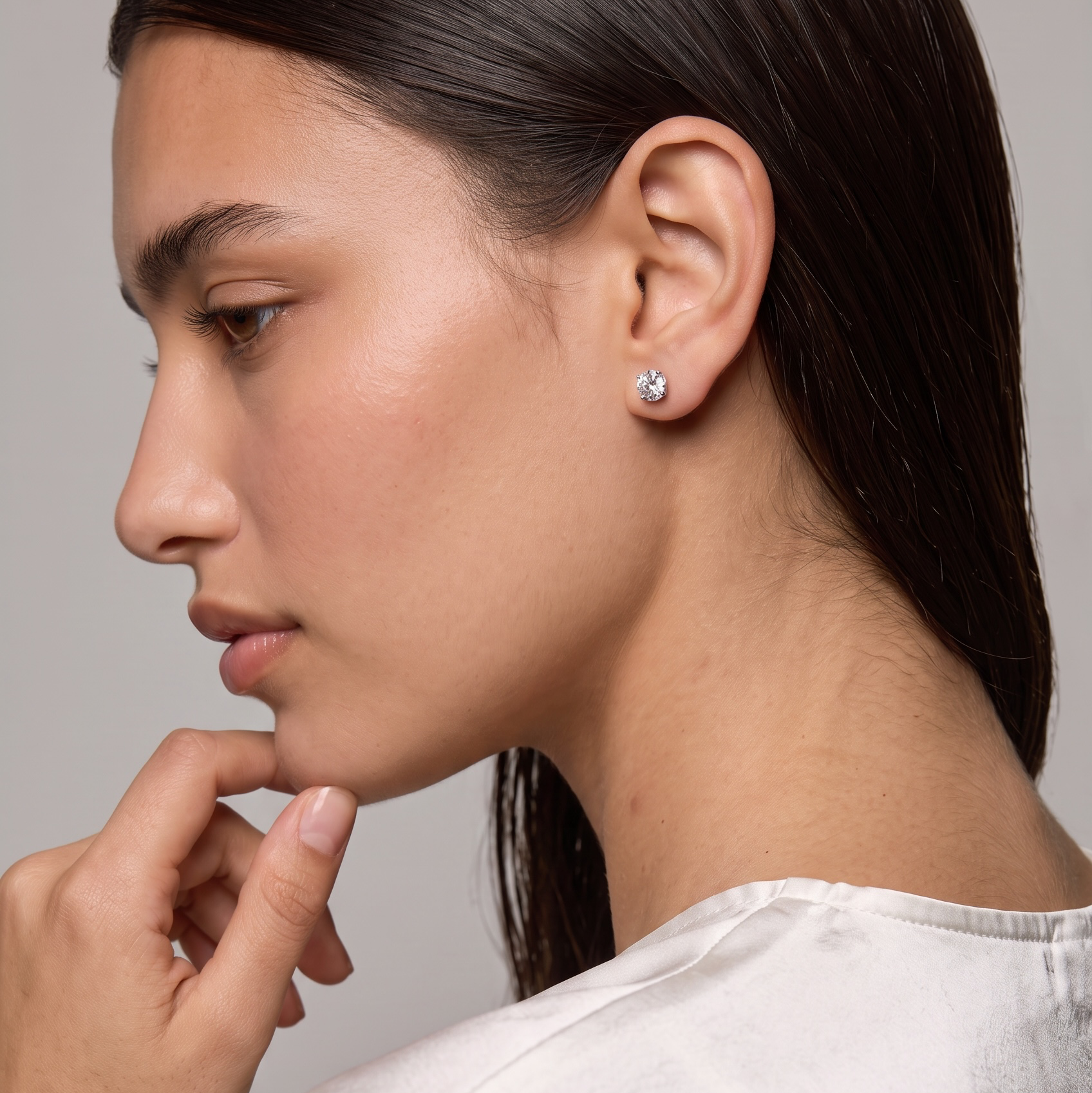 A close-up side profile of a woman wearing a small round clear gemstone stud earring, dressed in a white satin top against a neutral gray background.