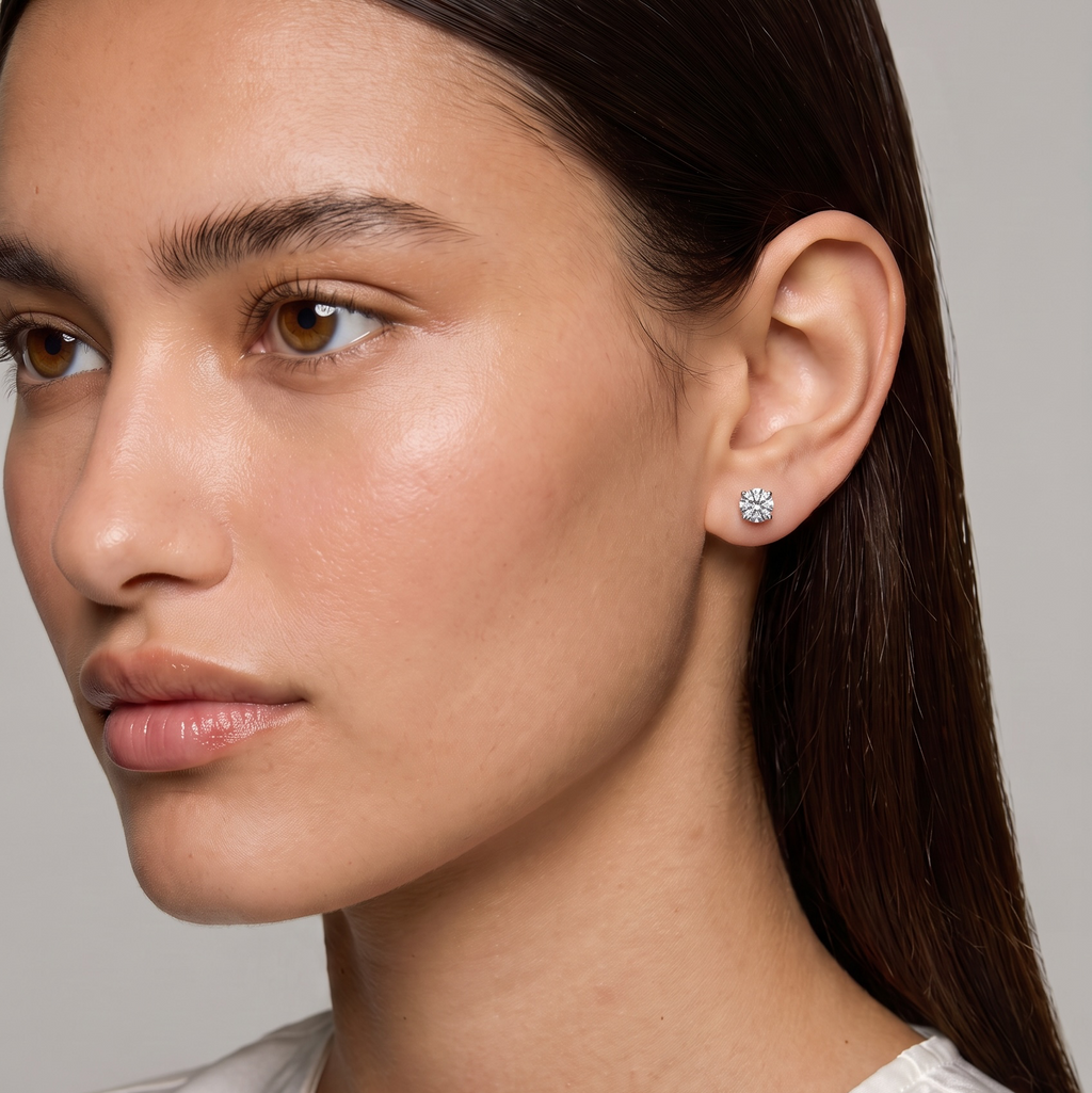 A close-up of a woman with dark hair wearing a small silver stud earring with a clear gemstone, set against a plain gray background.
