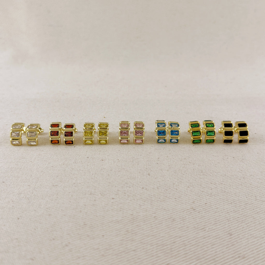 A row of nine gold-plated rectangular stud earrings with various colored rectangular gemstones, displayed on a light beige textured fabric background.