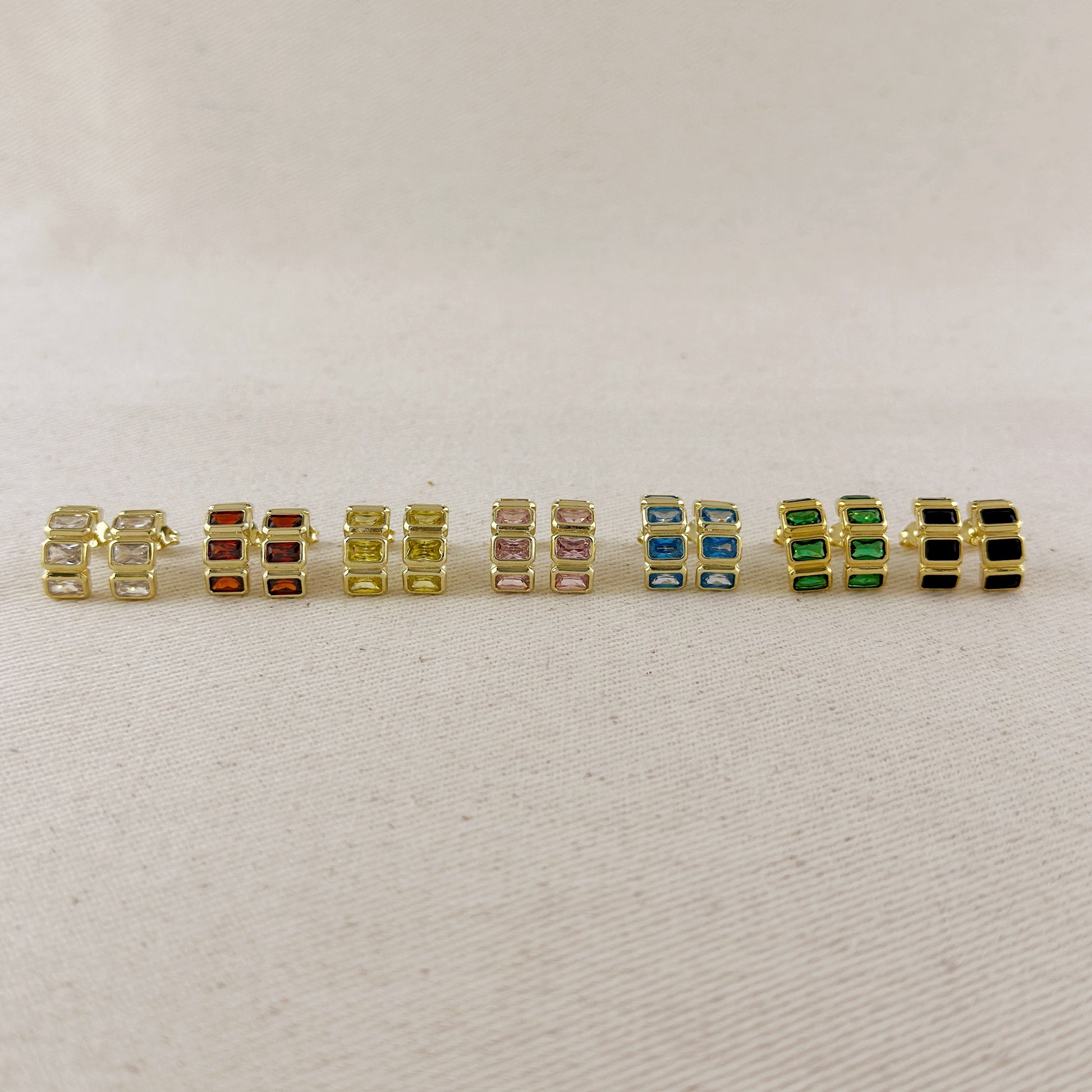 A row of nine gold-plated rectangular stud earrings with various colored rectangular gemstones, displayed on a light beige textured fabric background.