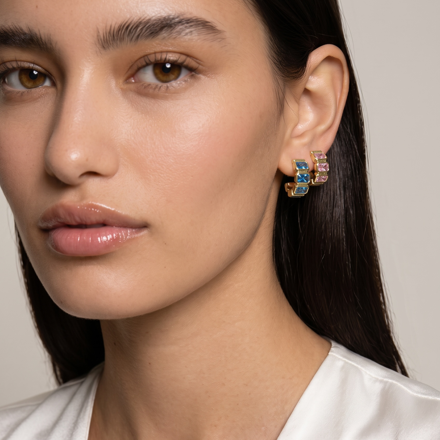 Close-up of a woman wearing a small gold hoop earring with colorful enamel details in blue, yellow, and orange, paired with a white satin top against a neutral beige background.