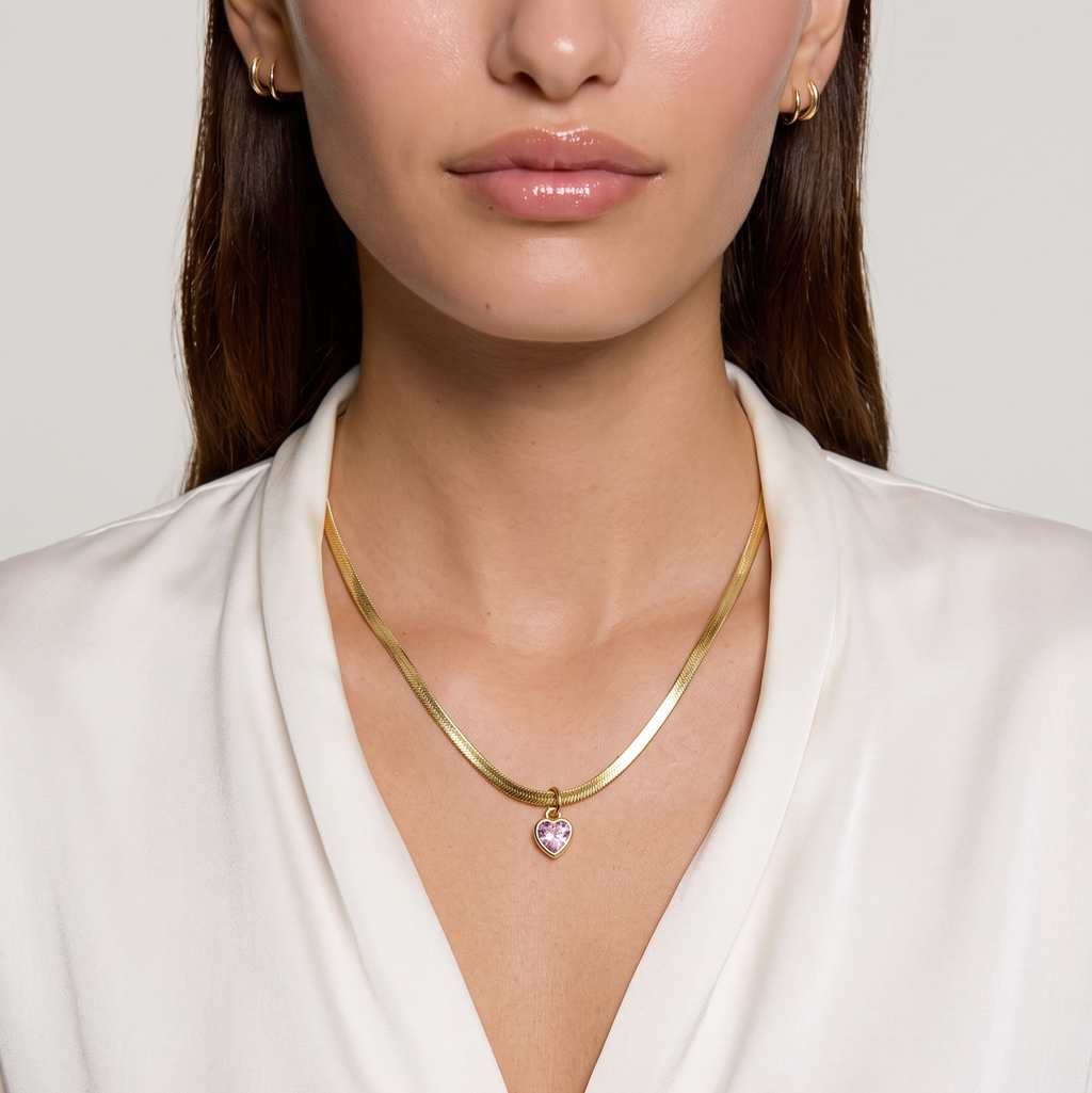 A close-up of a model wearing a delicate gold chain necklace with a small heart-shaped crystal pendant, paired with matching gold hoop earrings and a white satin blouse.