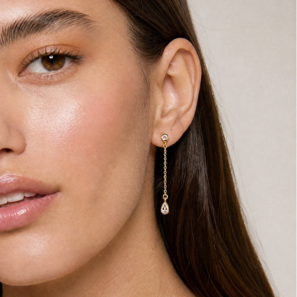 A close-up of a woman wearing a delicate gold dangle earring with a small clear teardrop gemstone, shown against a soft beige background.