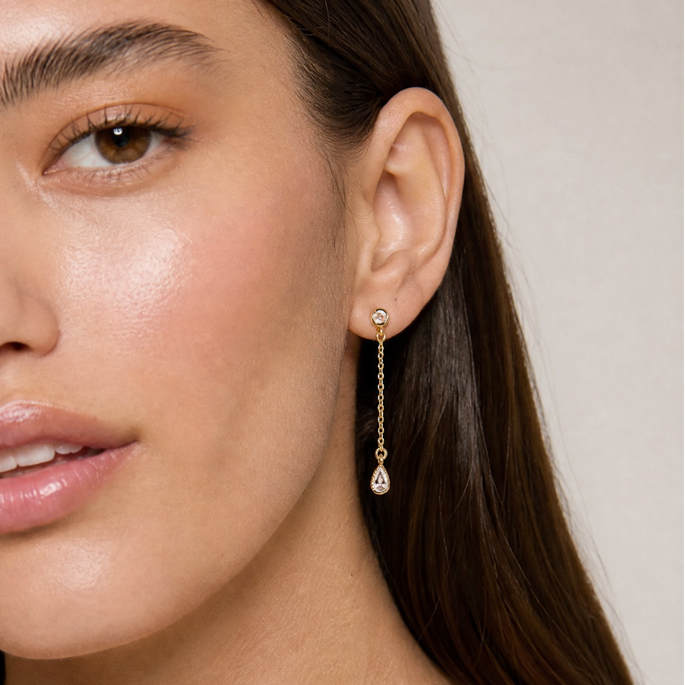 A close-up of a woman wearing a delicate gold dangle earring with a small clear teardrop gemstone, shown against a soft beige background.