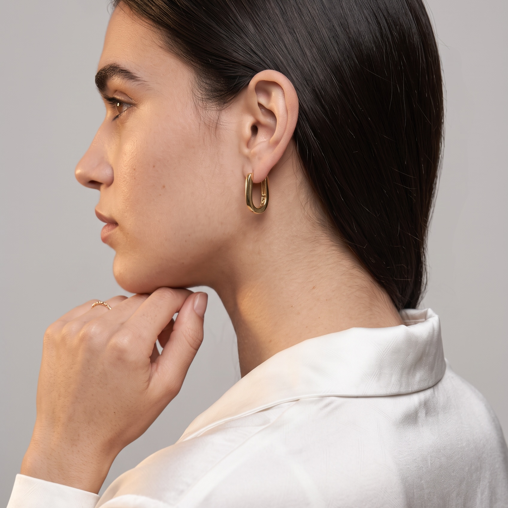 A woman in profile wearing a small gold hoop earring, shown from the back and side against a plain light gray studio background.