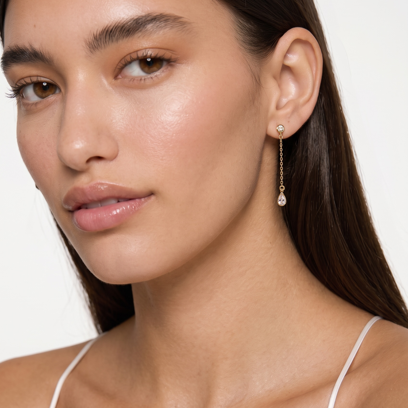 A close-up of a woman wearing a delicate gold dangle earring with a small clear stone, shown against a clean white background.
