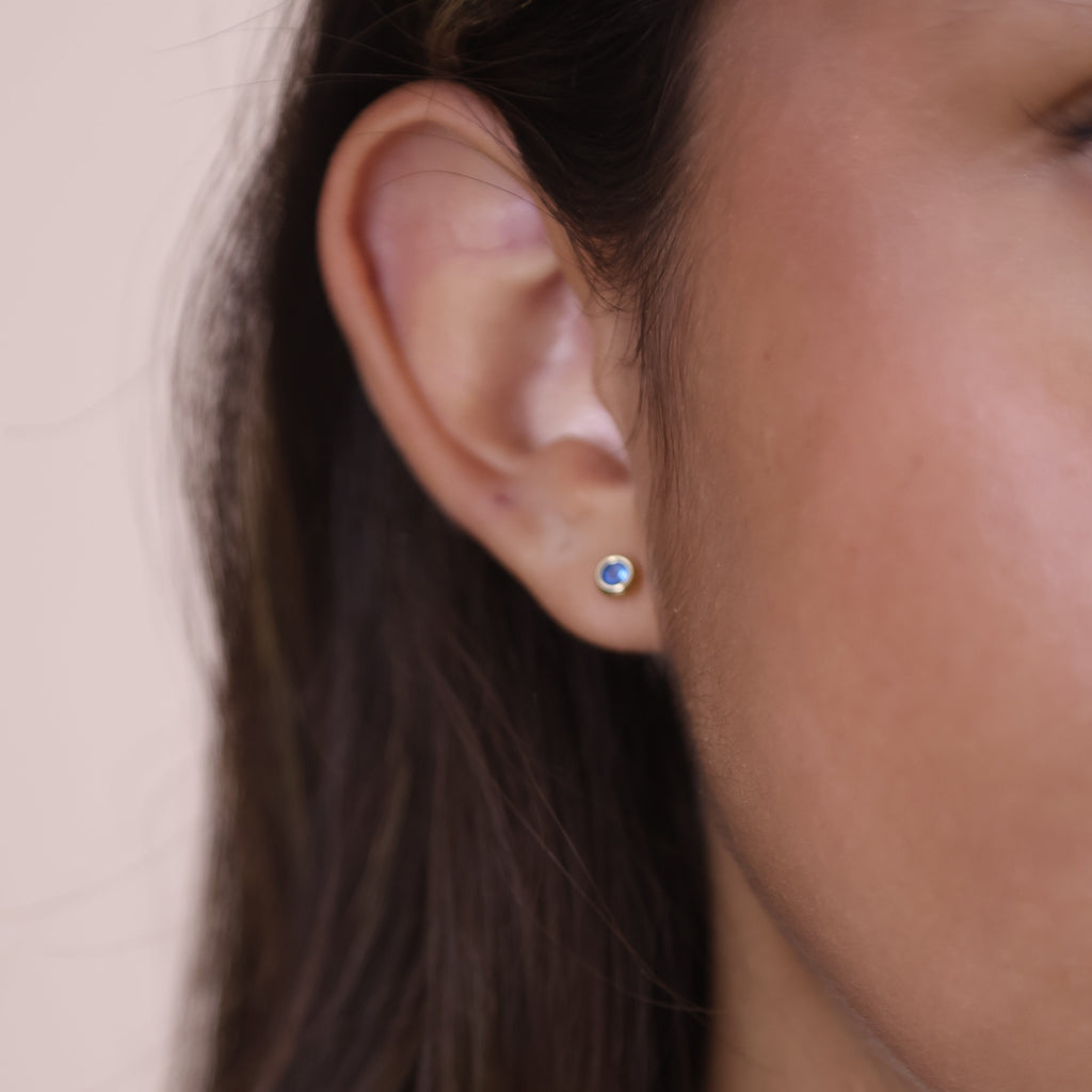 Close-up of dainty 18K gold filled stud earrings featuring a blue cubic zirconia birthstone on a woman's ear.