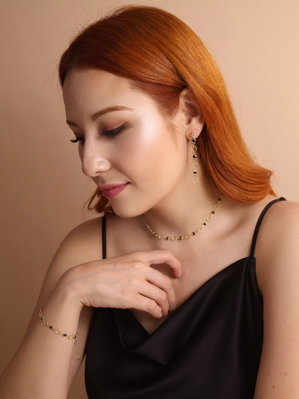 Woman with red hair wearing a black top and gold jewelry against a beige background